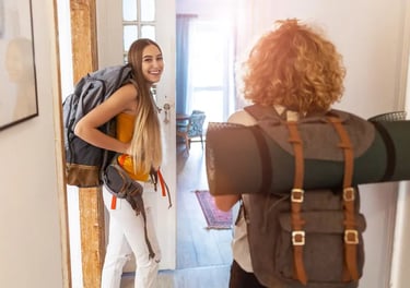 Two female backpackers with travel gear entering a bright hostel room for their vacation adventure.