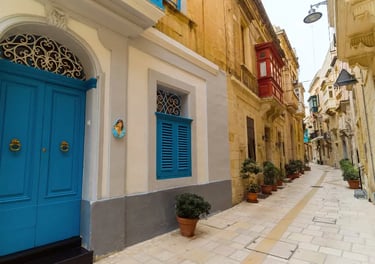 A narrow limestone street in Malta featuring traditional blue doors and red wooden balconies.