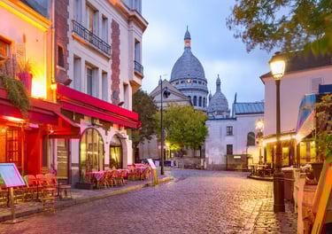 A charming cobblestone street in Montmartre, Paris, featuring local cafes and the Sacré-Cœur Basilica at dusk.