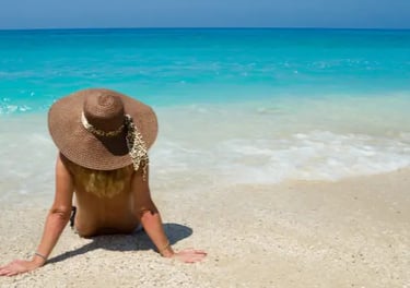A woman wearing a sun hat sitting on a tropical beach overlooking turquoise ocean water.