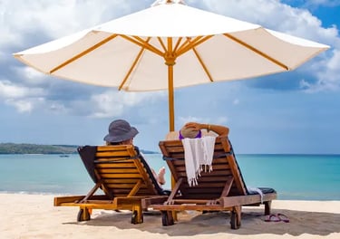 Two people relaxing on wooden lounge chairs under a white beach umbrella by the turquoise ocean.
