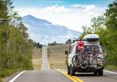 White camper van with mountain bikes driving on a scenic road toward distant mountains.