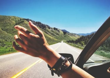 Hand reaching out a car window on a scenic summer road trip through green mountain hills.