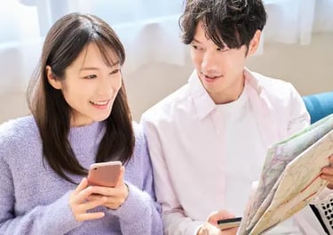 Smiling couple sitting on a sofa planning a trip using a smartphone and paper map.