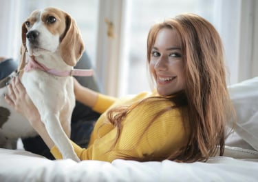 A smiling young woman in a yellow sweater relaxing on a bed with her pet beagle dog.