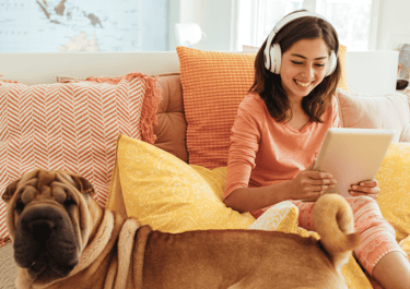A smiling young girl wearing headphones uses a digital tablet while sitting in bed with her Shar-Pei dog.