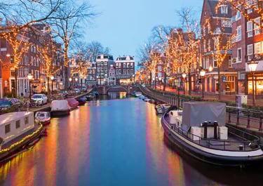 Amsterdam canal at dusk with festive Christmas lights on trees and historic Dutch architecture reflecting on the water.