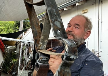 A bearded blacksmith holding a large custom metal sculpture with welded iron joints in an outdoor workshop.