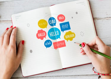 A person writing in an open notebook with colorful speech bubbles showing greetings in multiple languages.
