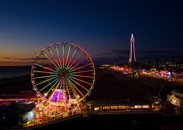 Illuminated Ferris wheel on Blackpool Central Pier at night with the Blackpool Tower in the background.