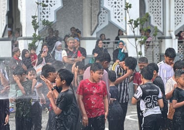 Young boys playing in water spray outside a mosque with traditional Islamic architecture.