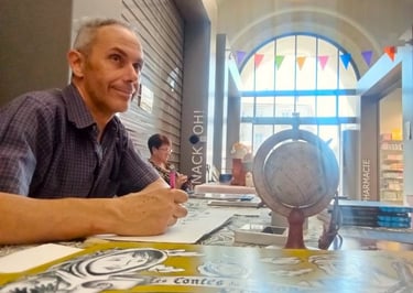Artist Dim signing books at a table with a globe and colorful bunting in a bright indoor market.