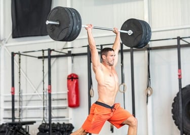 Shirtless man performing a barbell overhead press in a gym for strength training.