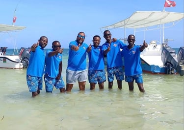 A group of smiling scuba intructors in blue shirts giving thumbs up in the ocean near boats.