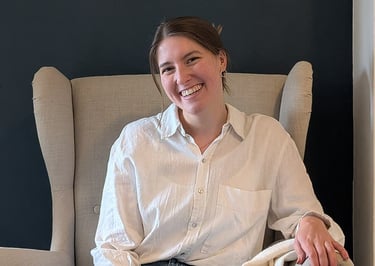 A smiling woman in a white linen shirt and jeans sits cross-legged in a beige wingback chair.