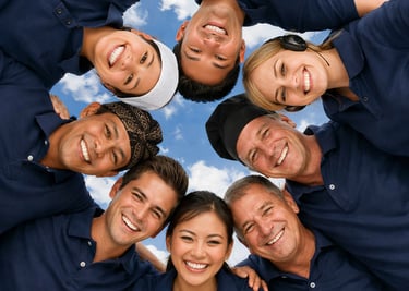 Diverse team of smiling coworkers in blue uniforms huddling in a circle under a blue sky.