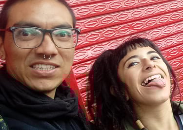 Un hombre sonriente con gafas y una mujer sacando la lengua, posando frente a una pared roja con tex