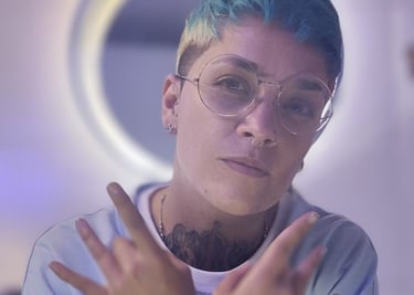 Persona con cabello corto azul y gafas, haciendo gestos con las manos frente a un aro de luz.