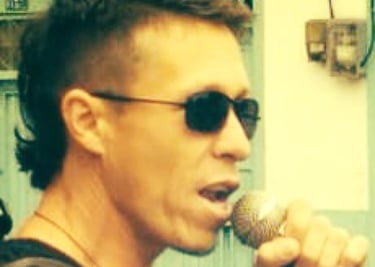 Un vocalista masculino con un corte de cabello mullet moderno y gafas de sol, cantando en un micrófo