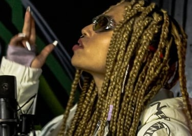 Artista con trenzas boxeadoras rubias, usando gafas de sol y una chaqueta acolchada blanca 