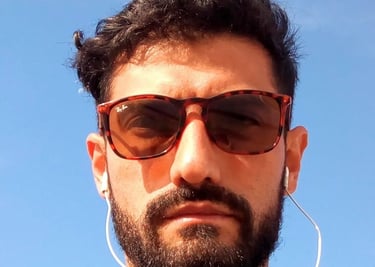 Hombre con barba usando gafas Ray-Ban de carey y audífonos blancos con cable, con el cielo azul 