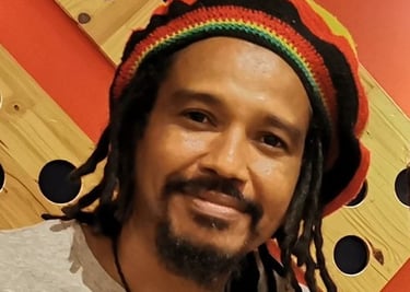 Un hombre con rastas, usando un gorro rasta colorido y una camisa gris, frente a una pared roja.