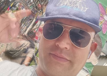 Un hombre con una gorra azul de béisbol y gafas de sol tipo aviador, posando para una selfie al aire