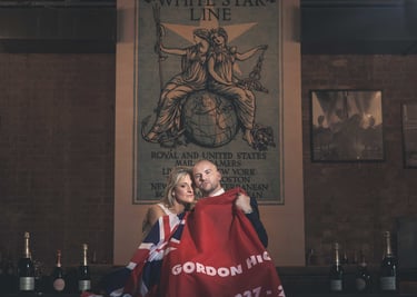 Couple holding a Union Jack flag under a vintage White Star Line poster in a brick room.