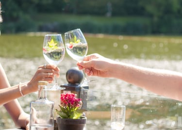 Zwei Hände stoßen mit Weingläsern im sonnigen Restaurant mit Seeblick an, warme Lichtstimmung
