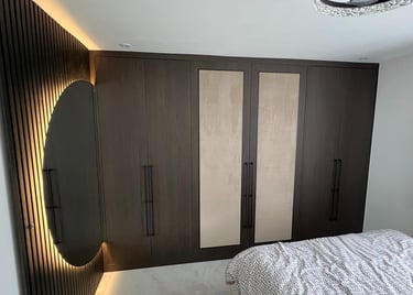 Modern dark wood fitted wardrobe with integrated lighting and a round backlit mirror in a bedroom.