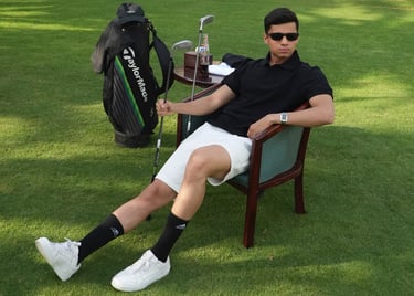 A young golfer wearing sunglasses relaxes in a chair on a green golf course with his TaylorMade bag and clubs.