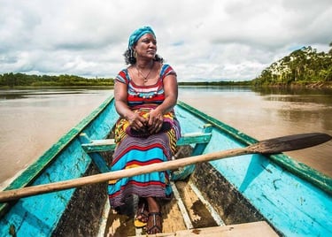 A woman in a colorful dress sitting in a wooden boat on a tropical river surrounded by rainforest.