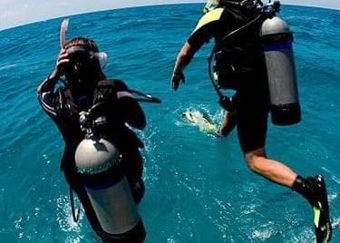 Two scuba divers jumping off a boat into clear blue ocean water with full dive gear.