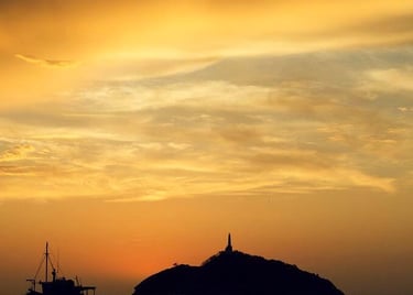 Silhouetted fishing boats on calm water at sunset with a lighthouse island on the horizon.