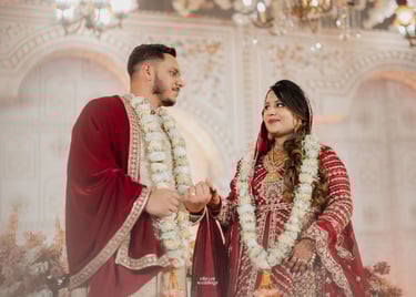 A couple in traditional red and white South Asian wedding attire posing under a chandelier.