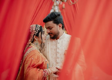 A couple in traditional Indian wedding attire posing behind red fabric curtains.