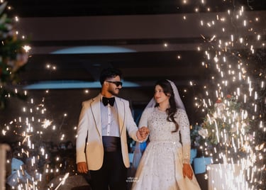 A bride and groom in a white tuxedo and sequined gown walk through indoor wedding sparklers.