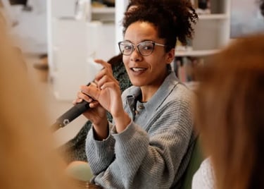 Una mujer con gafas y un suéter sentada hablando a un microfono