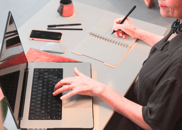 Uma mulher consulta o notebook aberto em cima da mesa enquanto faz anotações no caderno ao lado.