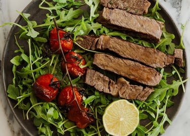 Sliced grilled steak served over fresh arugula with roasted cherry tomatoes and a lemon wedge.