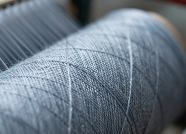 A macro photograph focusing on the intricate texture of Kota yarn. The thread is crisp and well-defined against a soft-focus background of the spinning machinery. Lighting highlights the metallic sheen of the fiber, incorporating #95A5BB tones.