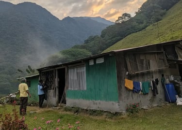 Rustic green wooden farmhouse in a tropical mountain valley with laundry hanging and misty peaks.