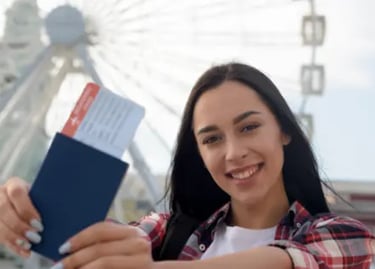 A smiling young woman holding a passport and flight boarding pass in front of a white ferris wheel.