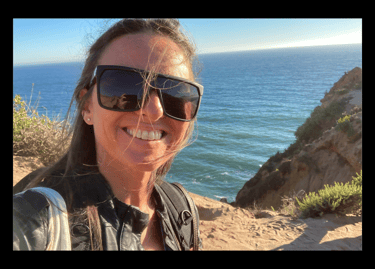 Michael Alexa Pitts on a rocky Pacific coastline with ocean behind, smiling, with black sunglasses & strands of hair in face