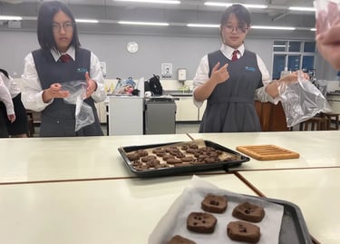 兩位穿著校服的學生站在廚藝教室中，手持食材袋，在鋪有新鮮出爐朱古力曲奇的檯前交流，場景展現學習動手能力與課堂合作精神。