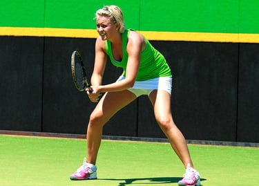 A blonde female tennis player in a green top and white skirt waits to return a serve on a grass court.