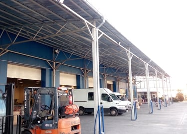 Logistics warehouse loading bay with parked forklifts, delivery vans, and a metal canopy roof.