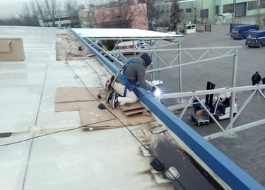 Professional welder with safety harness welding a steel roof structure on an industrial building site.