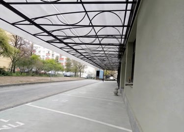Modern polycarbonate canopy with decorative black metal frame over a sidewalk and parking area.