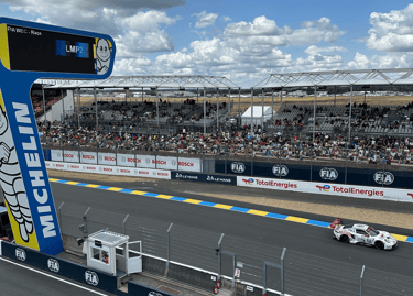 A white Porsche race car speeds past the grandstands at the 24 Hours of Le Mans circuit under a cloudy sky.